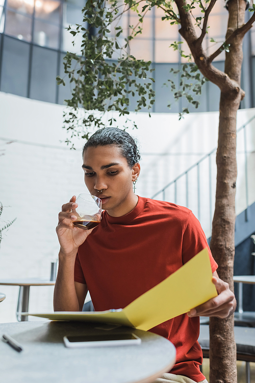 Young african american man holding glass of coffee and paper folder near smartphone in cafe