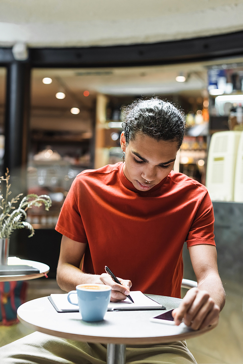 African american man writing on notebook near coffee and smartphone in cafe