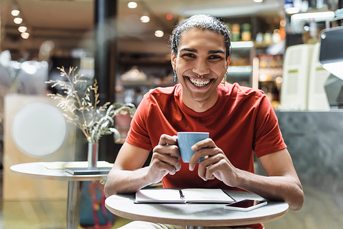Cheerful african american man smiling at camera while holding coffee near notebook in cafe