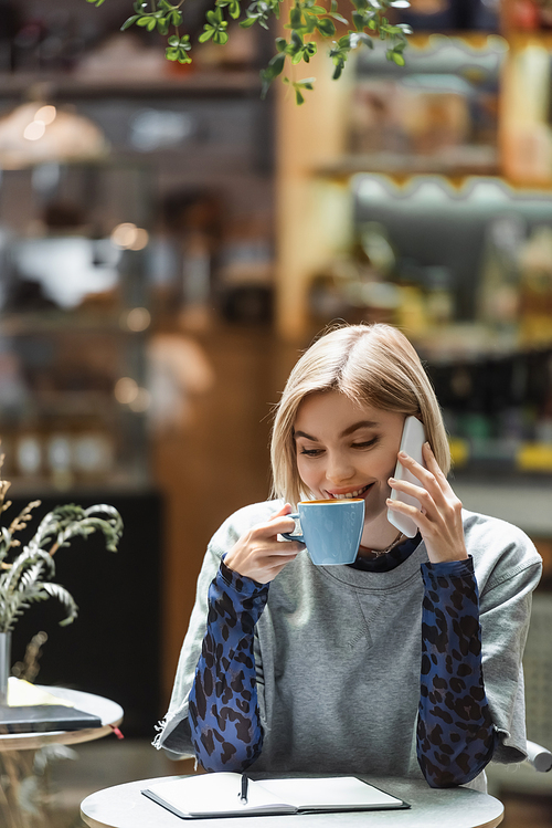 Positive woman talking on smartphone and drinking coffee near notebook in cafe
