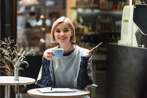 Smiling blonde woman holding cellphone and coffee near notebook in cafe