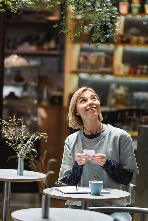 Smiling blonde woman using smartphone and looking up near coffee and notebook in cafe