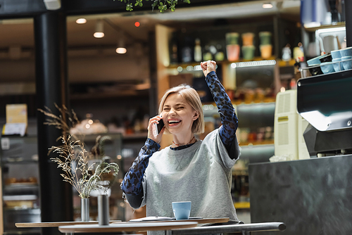 Excited blonde woman talking on smartphone near coffee and notebook in cafe