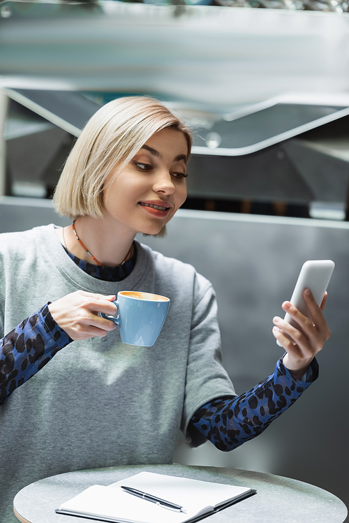 Young blonde woman holding cup and using smartphone near notebook in cafe