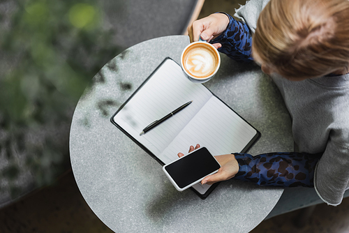 Overhead view of blonde woman using smartphone and holding cup of coffee near notebook in cafe