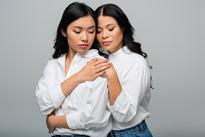 brunette mother and asian young daughter hugging each other and holding hands isolated on grey