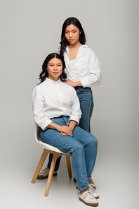 full length of asian mother sitting on chair near daughter in blue jeans and white shirt on grey