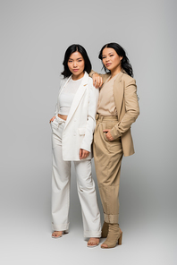 full length of trendy asian mother and young adult daughter in suits posing on grey