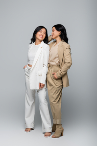 full length of stylish asian mother and young adult daughter in suits smiling on grey
