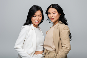 stylish asian mother and smiling young adult daughter in suit isolated on grey