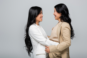 stylish asian mother and happy young adult daughter in suit hugging isolated on grey