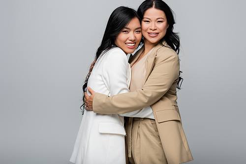 cheerful asian mother and happy young adult daughter in suit hugging isolated on grey