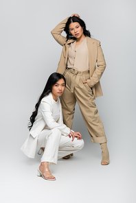 full length of stylish asian mother in beige suit and trendy young adult daughter posing on grey