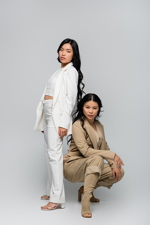 full length of trendy asian mother in beige suit and stylish young adult daughter posing on grey