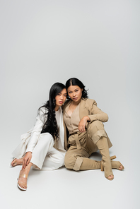 full length of stylish asian mother in beige suit and brunette young adult daughter sitting on grey