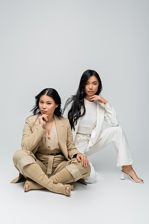 full length of stylish asian mother and brunette young adult daughter sitting on grey