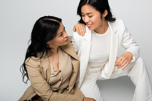 stylish asian mother and happy young adult daughter looking at each other on grey