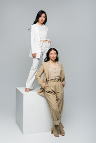 full length of asian young adult woman standing on cube near brunette mother posing on grey