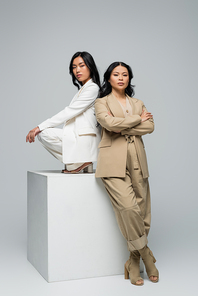 full length of asian young adult woman sitting on cube near brunette mother posing on grey