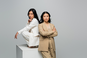 asian young adult woman sitting on cube near brunette mother posing with crossed arms isolated on grey