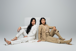full length of asian young adult woman sitting with brunette mother near cube on grey