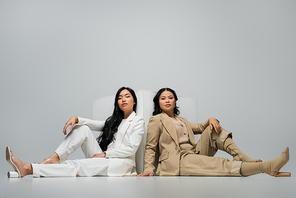full length of trendy asian young adult woman sitting with brunette mother near cube on grey