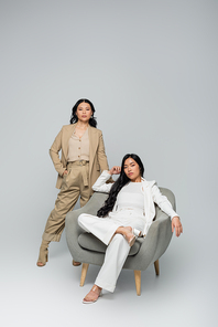 full length of young adult asian woman sitting in armchair near stylish mother on grey