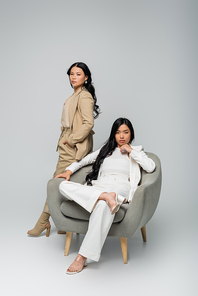 full length of young adult asian woman sitting in armchair near trendy mother on grey