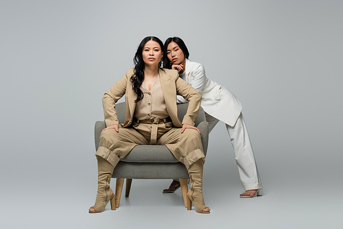 full length of asian woman sitting in armchair near trendy young daughter on grey