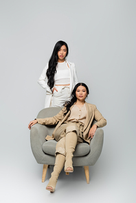 full length of asian woman sitting in armchair near trendy young daughter standing with hand in pocket on grey