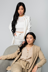 asian woman sitting in armchair near trendy young daughter standing with hand in pocket isolated on grey