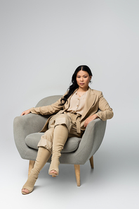 full length of trendy asian woman sitting in armchair on grey