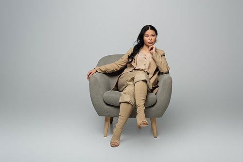 full length of stylish asian woman sitting in armchair on grey
