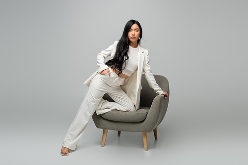 full length of young asian woman leaning on armchair while posing with hand in pocket on grey