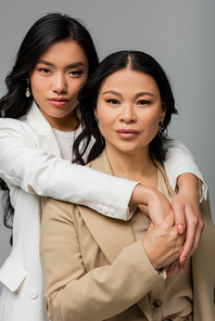young asian daughter hugging brunette mother in beige suit isolated on gray
