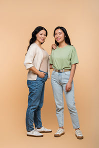full length of happy asian mother in jeans standing with hand in pocket near daughter on beige