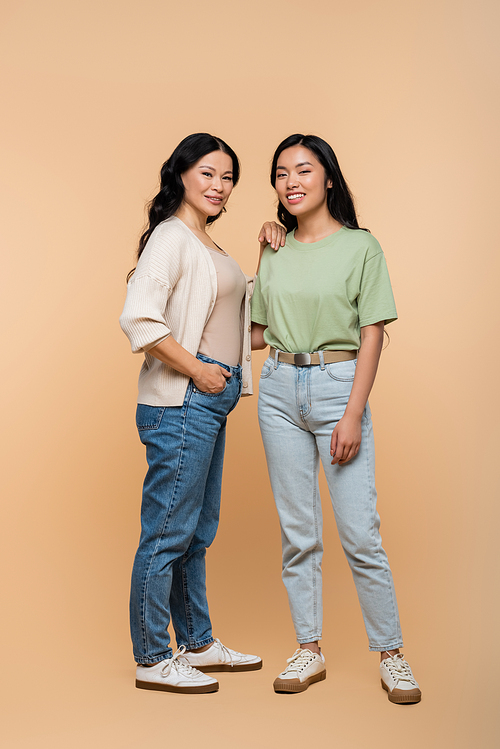 full length of happy asian mother in jeans standing with hand in pocket near daughter on beige
