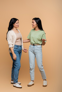 full length of happy asian mother in jeans looking at daughter on beige