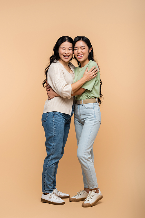 full length of cheerful asian mother hugging young adult daughter on beige