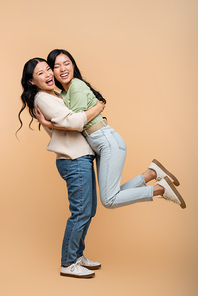 full length of cheerful asian mother lifting daughter on beige