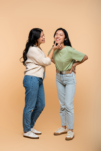 full length of cheerful asian mother touching face of pleased young adult daughter on beige