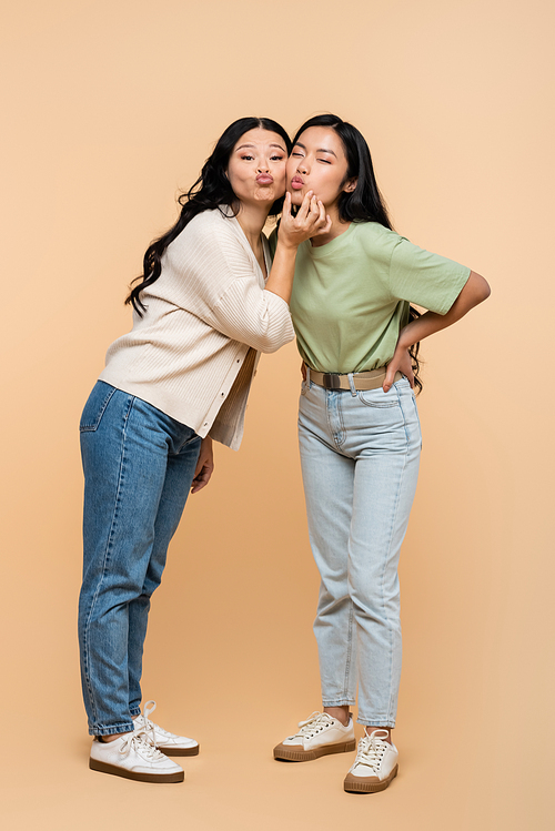 full length of funny asian mother touching face of young adult daughter and pouting lips on beige