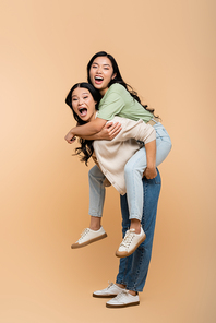 full length of asian mother piggybacking adult daughter with open mouth on beige