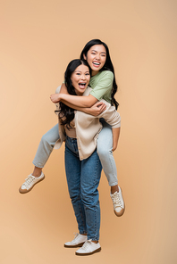 full length of amazed asian mother piggybacking adult daughter with open mouth on beige