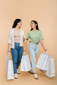 full length of asian mother and happy young adult daughter smiling while holding shopping bags on beige