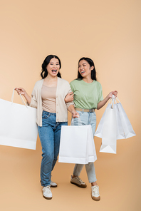 full length of asian mother and young adult daughter smiling while walking with shopping bags on beige