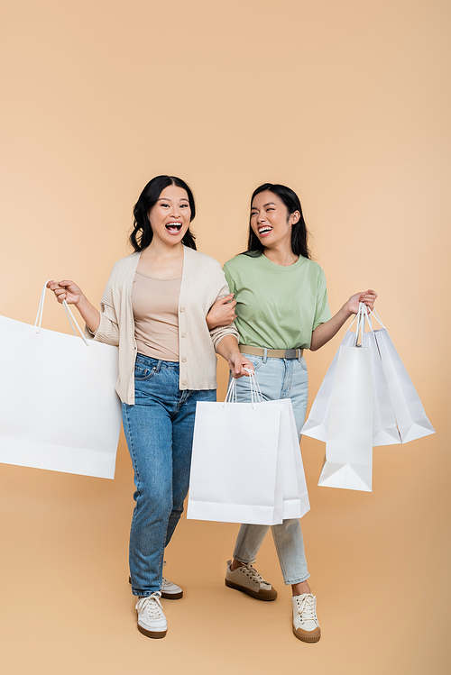 full length of asian mother and young adult daughter smiling while walking with shopping bags on beige