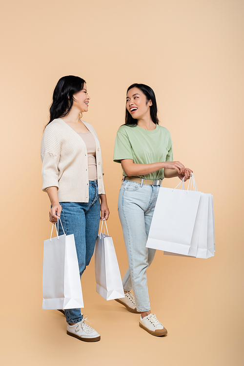 full length of asian mother and young adult daughter smiling while holding shopping bags on beige