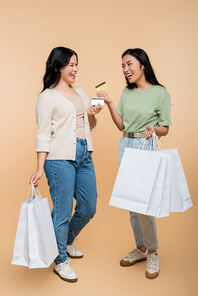 full length of happy asian mother and daughter with shopping bags and credit cards on beige