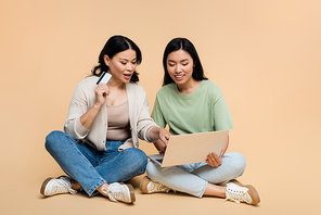 asian mother and adult daughter with credit card sitting with laptop during online shopping on beige